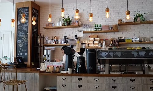 Interiér kavárny — barový pult s profesionálním kávovarem a mlýnkem, nad ním závěsná světla a police s konvicemi, za pultem barista